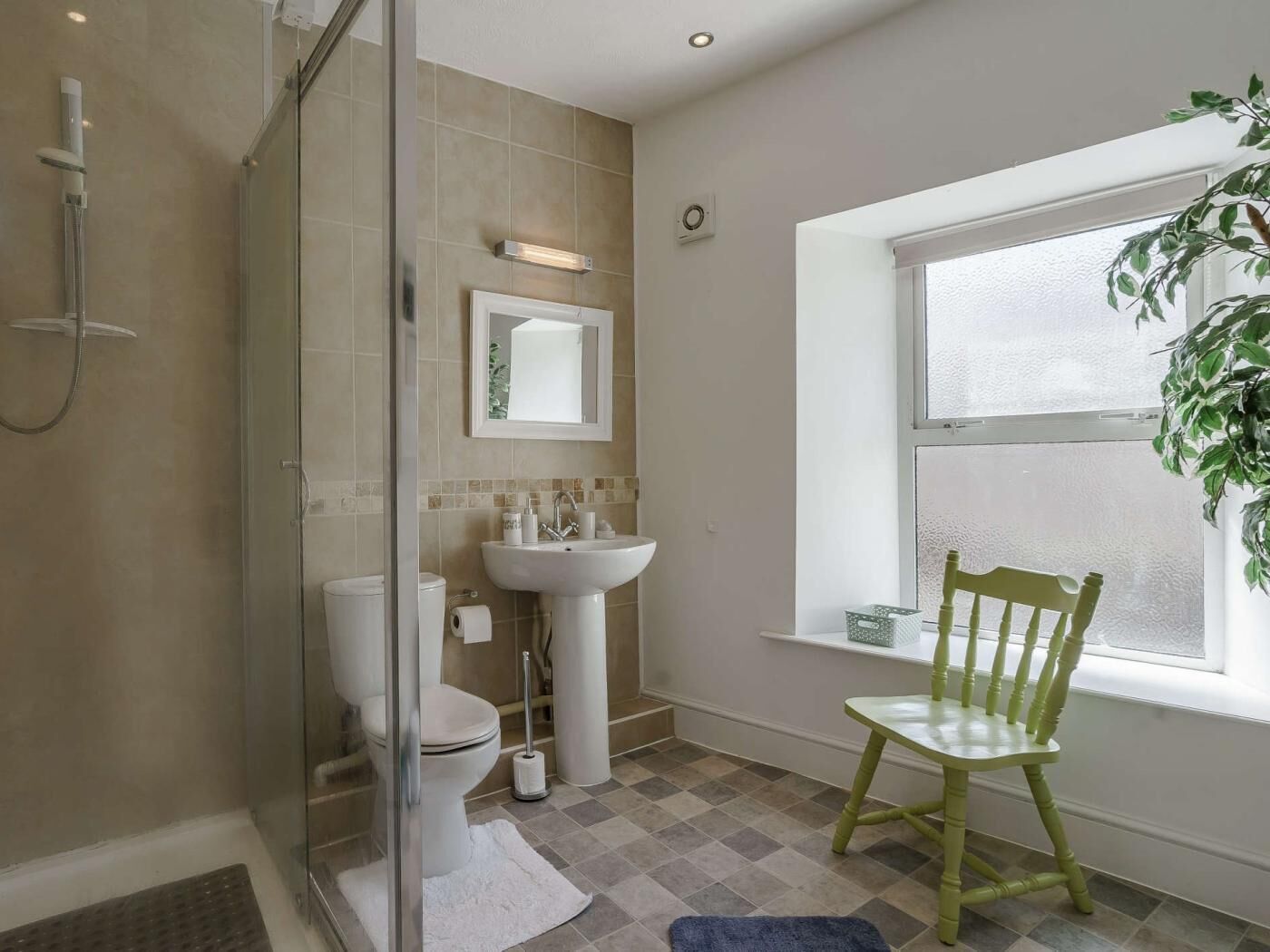 Une salle de bain avec une douche à parois de verre, des toilettes, un lavabo et une chaise près d’une fenêtre.
