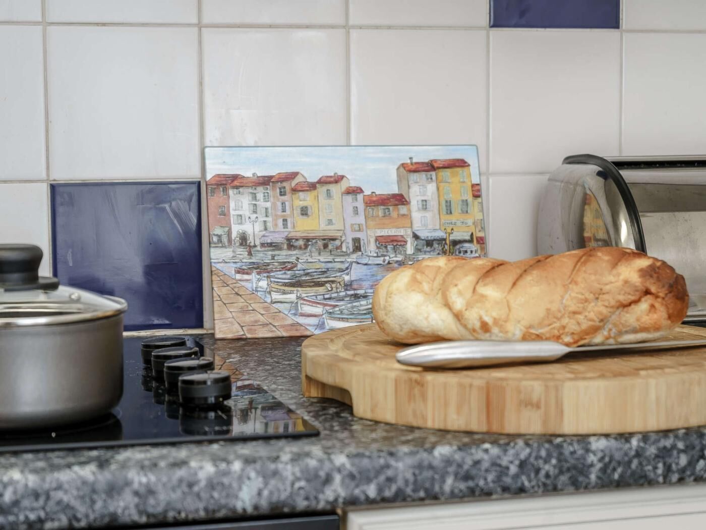 A kitchen with a loaf of bread on a cutting board, a pot on the stove, and a colorful street scene painting.
