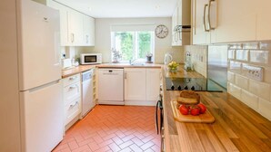 Eine moderne Küche mit weißen Schränken, einer Mikrowelle, einem Geschirrspüler, einem Kühlschrank, einem Fenster mit Blick auf Grünflächen und einer Holz Arbeitsplatte mit Schneidebrett und Tomaten.