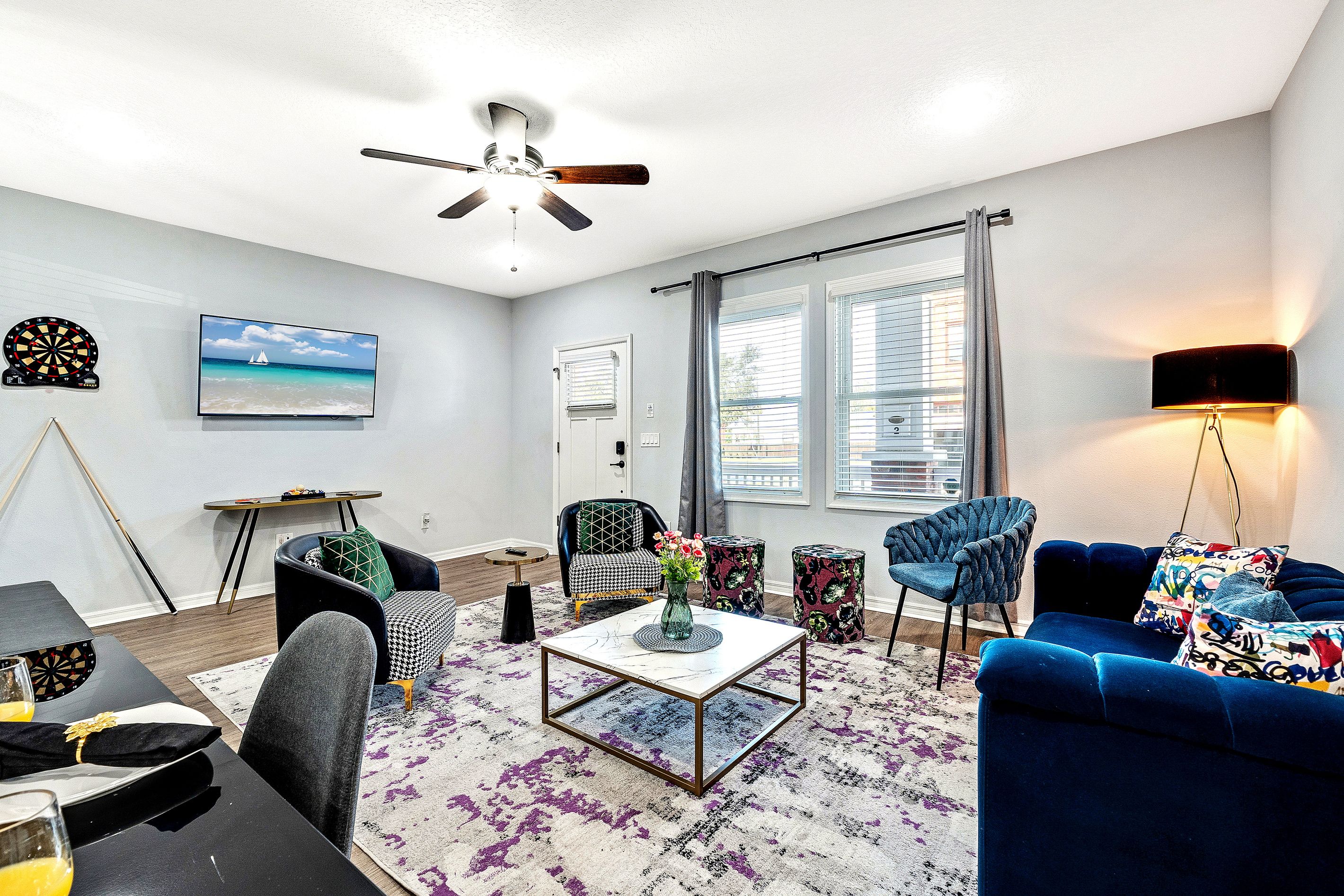 A modern living room with a ceiling fan, a flat-screen TV, a dartboard, a coffee table, and a blue sofa.