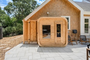 A wooden sauna structure with a door, a window, and a small outdoor seating area with chairs. - New Construction Designer Home: Private Outdoor Oasis, Hot Tub, Sauna (Eastham)