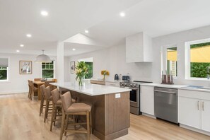 A modern kitchen with a central island, bar stools, stainless steel appliances, and large windows. - New Construction Designer Home: Private Outdoor Oasis, Hot Tub, Sauna (Eastham)