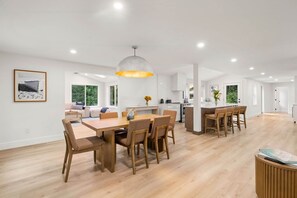 A spacious living area with a dining table and chairs, a kitchen in the background, and large windows offering a view of the outdoors. - New Construction Designer Home: Private Outdoor Oasis, Hot Tub, Sauna (Eastham)