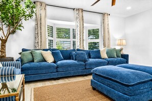 A living room with a blue sofa set, a coffee table, a lamp, and a large window with curtains. - Family House | Fenced Pool, Game Room, Playset | 5BR | 14 Beds (Clearwater)
