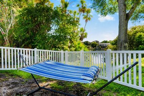 A hammock with a striped fabric, positioned in front of a white picket fence, surrounded by lush greenery and trees. - Family House | Fenced Pool, Game Room, Playset | 5BR | 14 Beds (Clearwater)