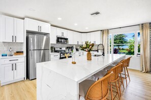 A modern kitchen with white cabinetry, stainless steel appliances, a marble island, and a view of greenery outside. - Family House | Fenced Pool, Game Room, Playset | 5BR | 14 Beds (Clearwater)