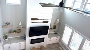 Una sala de estar moderna con televisor de pantalla plana, estanterías de madera, ventilador de techo y un amplio ventanal.