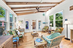 A spacious room with wooden flooring, wicker furniture, and large windows offering views of the outdoors. - One-of-a-Kind Artist Retreat, 4Min Walk to Beach (Corolla)