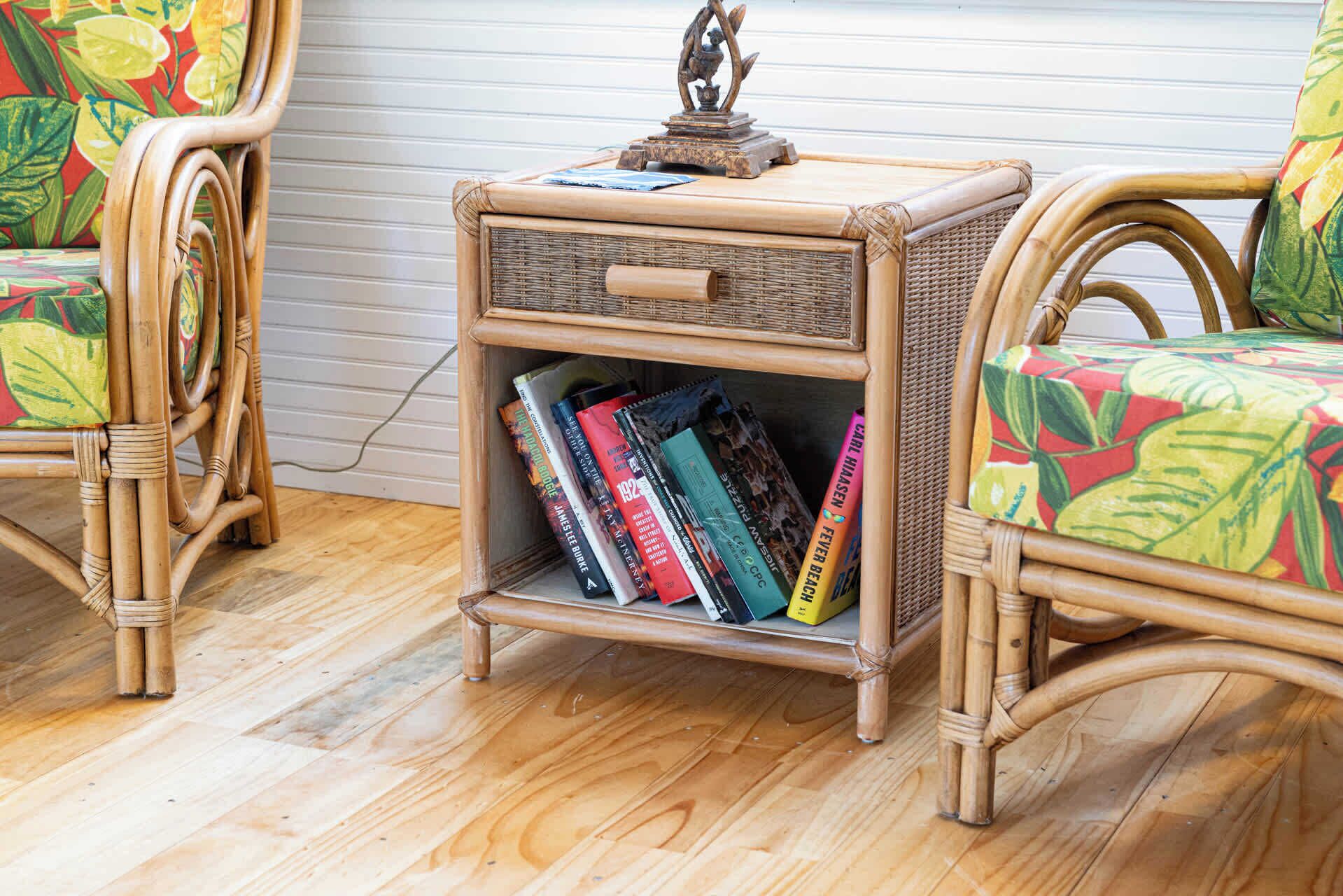 Une table de chevet en osier, dotée d’un tiroir et d’une étagère pour les livres, placée entre deux fauteuils en osier à motifs floraux.