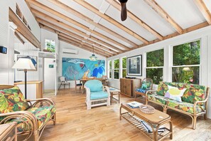 A spacious room with wooden beams, a large painting, a television, and a sofa with a tropical print. - One-of-a-Kind Artist Retreat, 4Min Walk to Beach (Corolla)