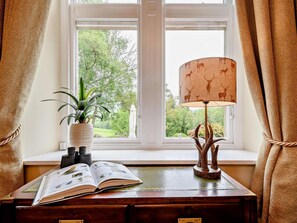 Ein Fenster mit Blick auf Grünflächen, eine Lampe mit Hirschmuster, eine Topfpflanze und ein offenes Buch auf einem Holztisch.