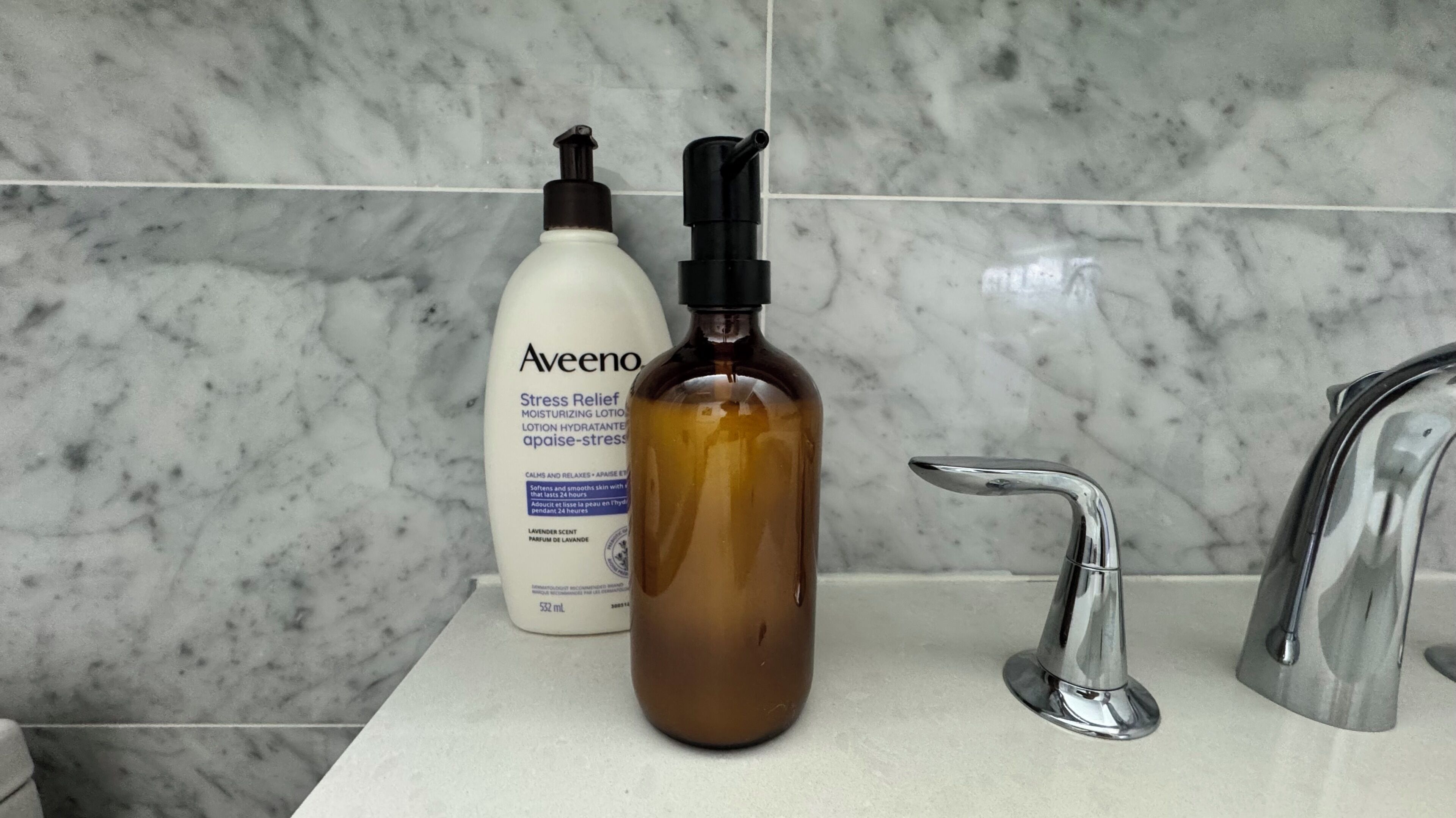 Une salle de bain avec un pare-baignoire en marbre, un flacon blanc de lotion Aveeno et un flacon marron doté d’un distributeur à pompe.