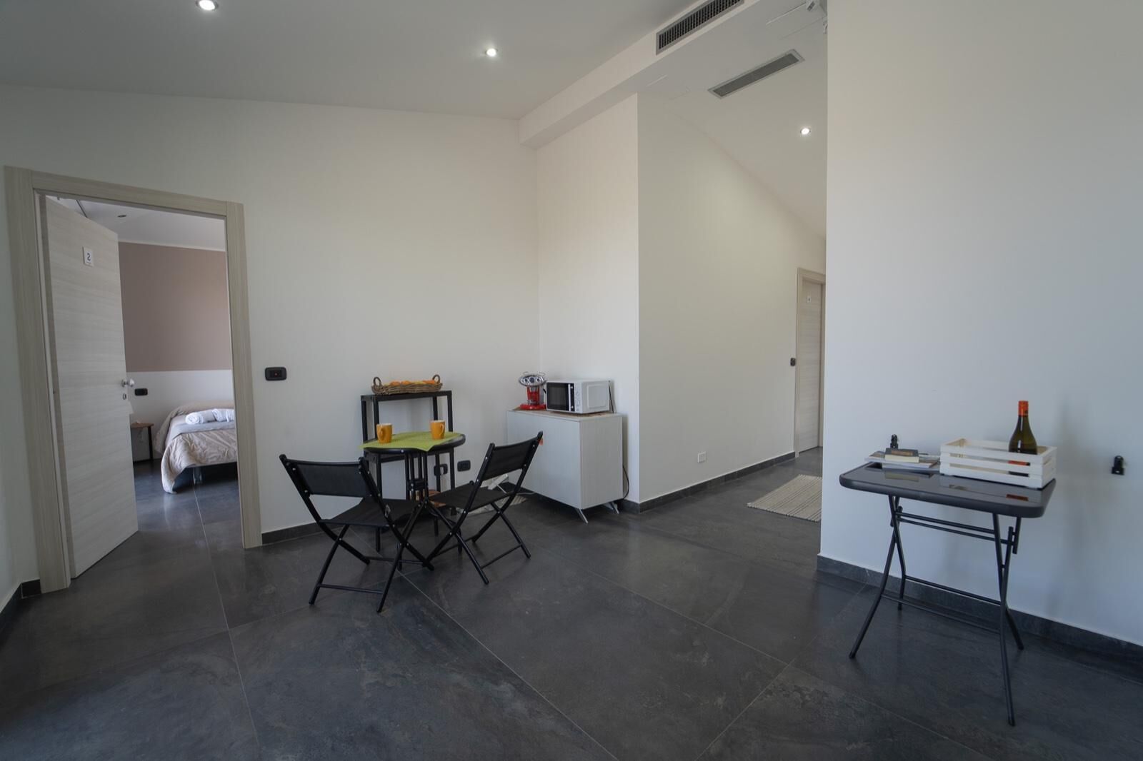 A modern hotel room with a bed, a dining table with chairs, a small kitchenette, and a desk with a chair.