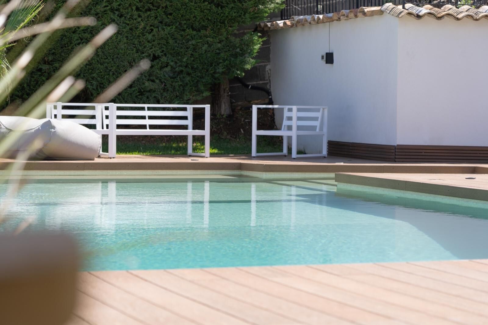 Une piscine entourée d’une clôture blanche, avec une chaise blanche à proximité et un bâtiment blanc en arrière-plan.