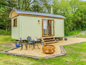 A small, single-story cabin with a wooden door and windows, a small patio with a table and chairs, and a fire pit. - 1 Bed in Pembroke (oc-83421) (Lamphey)
