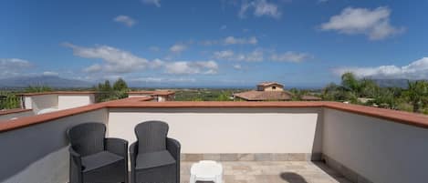 A rooftop terrace with two wicker chairs and a small white stool, a tiled floor, and a view of distant buildings and greenery.