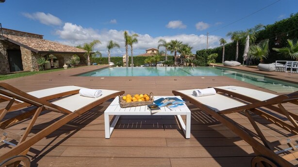 A poolside area with lounge chairs, a table with magazines and fruit, and a view of a pool and distant buildings. - Camera Luna Perla - Partinico (Monreale)