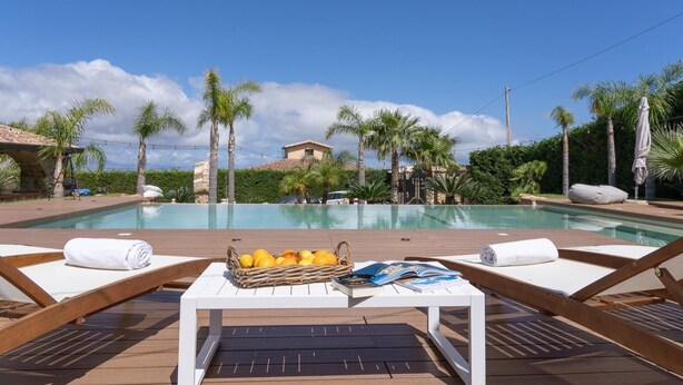 Zona junto a la piscina con sillones de descanso, una mesa con una cesta de fruta y vista a un edificio de fondo.
