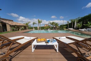 A poolside area with lounge chairs, a table with magazines and fruit, and a view of a pool and distant buildings. - Camera Luna Serena - Partinico (Monreale)