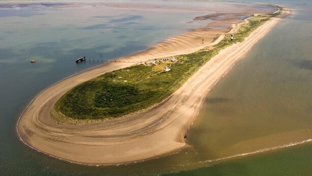 Vista aérea de una zona costera con una playa arenosa curva, un pequeño asentamiento con edificios y un faro.
