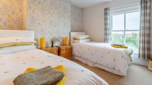 Un dormitorio con dos camas, una ventana con cortinas, una mesita de noche con lámpara y un papel tapiz con motivos florales.