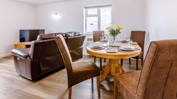 Een woonkamer met een bank, stoelen, een eettafel voor vier personen, een televisie en een raam.