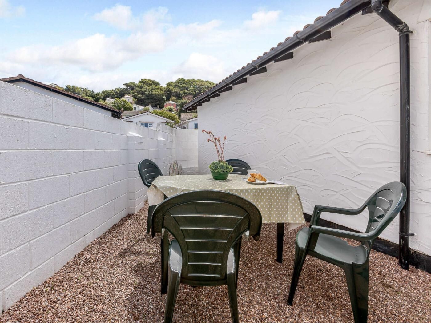 A small outdoor patio with a table and chairs, a white wall, and a view of a residential area.