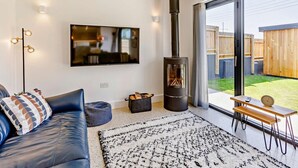 Ein modernes Wohnzimmer mit großem Fernseher, einem schwarzen Kaminofen, einer Couch und Blick auf einen Garten.