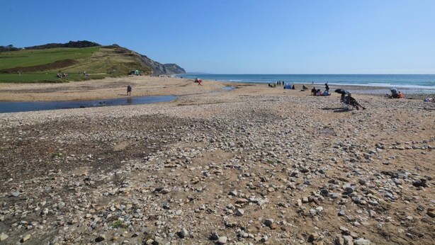 A pebbly beach with tents and people, a grassy hill in the background, and a clear sky. - 5 Bed in Bridport (oc-83584) (Charmouth)