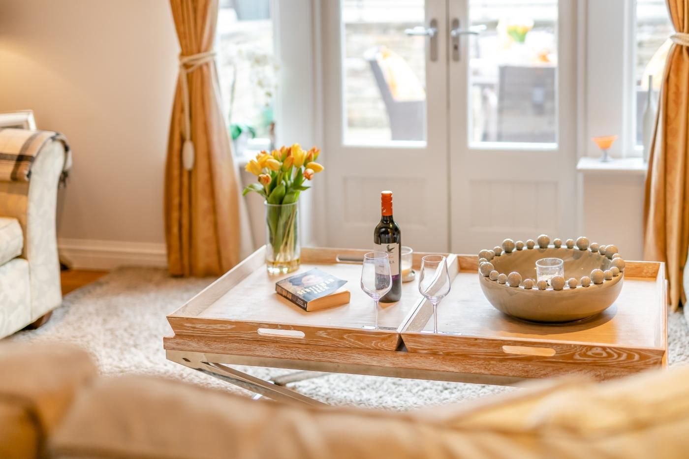 Una bandeja con una botella de vino, copas, un libro, un florero con flores y un cuenco decorativo sobre una mesa de madera en una habitación con vistas a través de una puerta de cristal.