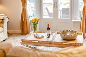 Una bandeja con una botella de vino, copas, un libro, un florero con flores y un cuenco decorativo sobre una mesa de madera en una habitación con vistas a través de una puerta de cristal.