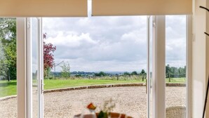 A dining area with a table set for two, large windows, and a view of a gravel field and trees. - 1 Bed in Corsham (oc-83690) (Box)