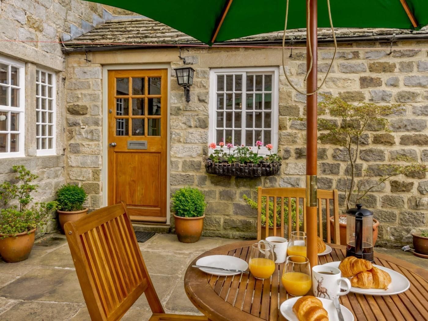 A stone building with a wooden door, windows, and a table set for breakfast with plates, glasses, and a coffee pot.