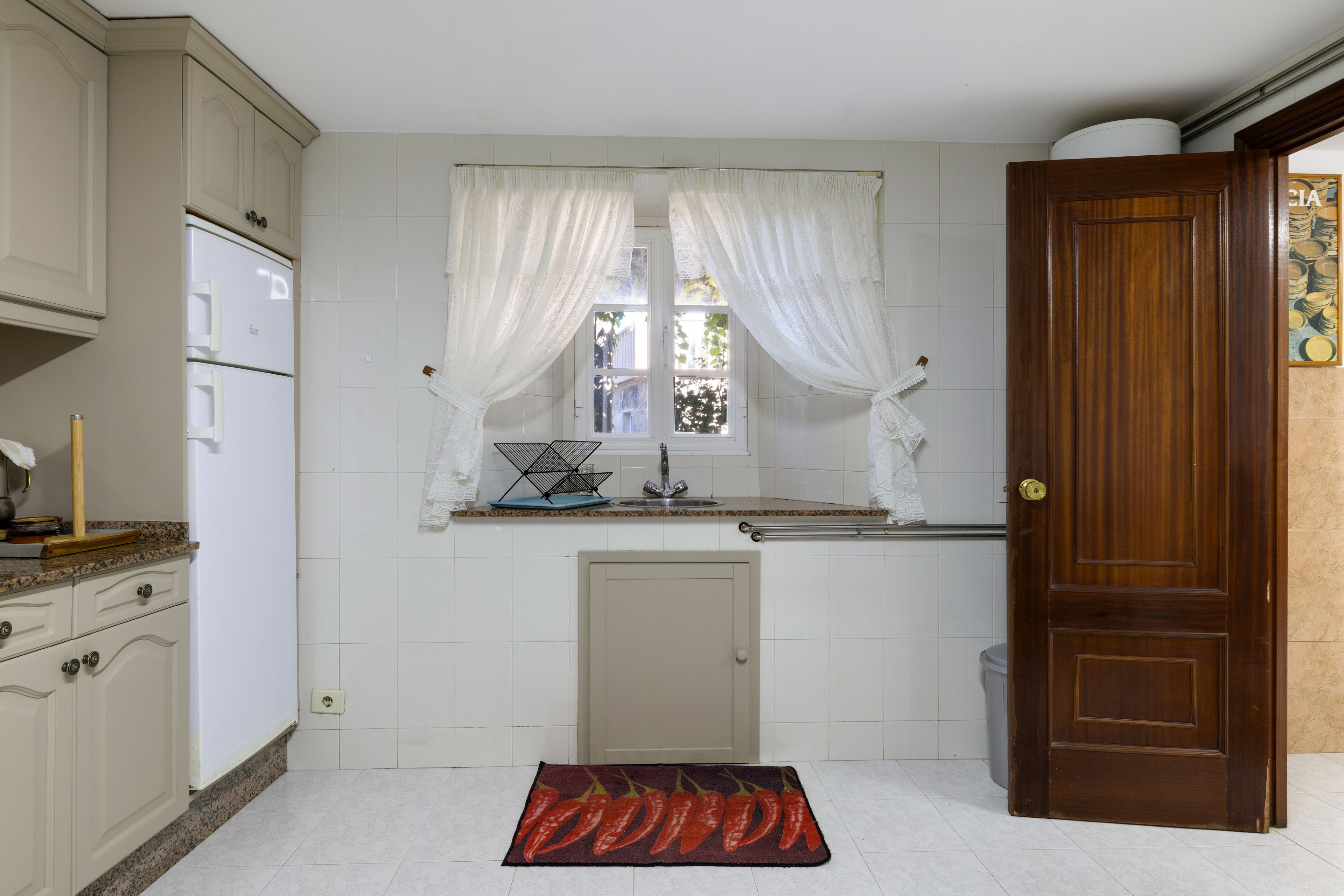 A kitchen with white cabinets, a window with sheer curtains, a wooden door, and a kitchen mat.