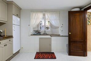 A kitchen with white cabinets, a window with sheer curtains, a wooden door, and a kitchen mat.