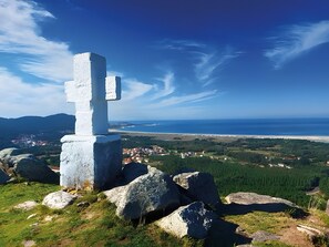 A white cross monument surrounded by rocks and greenery, overlooking a coastal town and the sea.