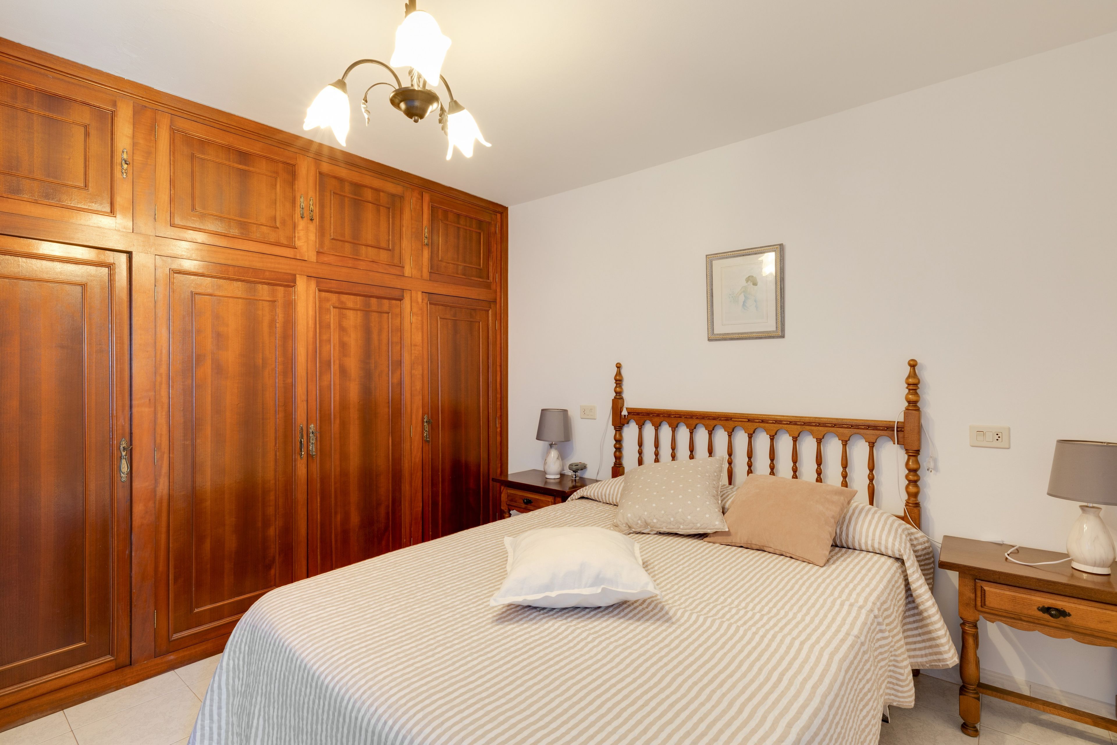 Une chambre avec une grande armoire, un lit avec une literie à rayures, une table de chevet avec une lampe et un tableau encadré au mur.