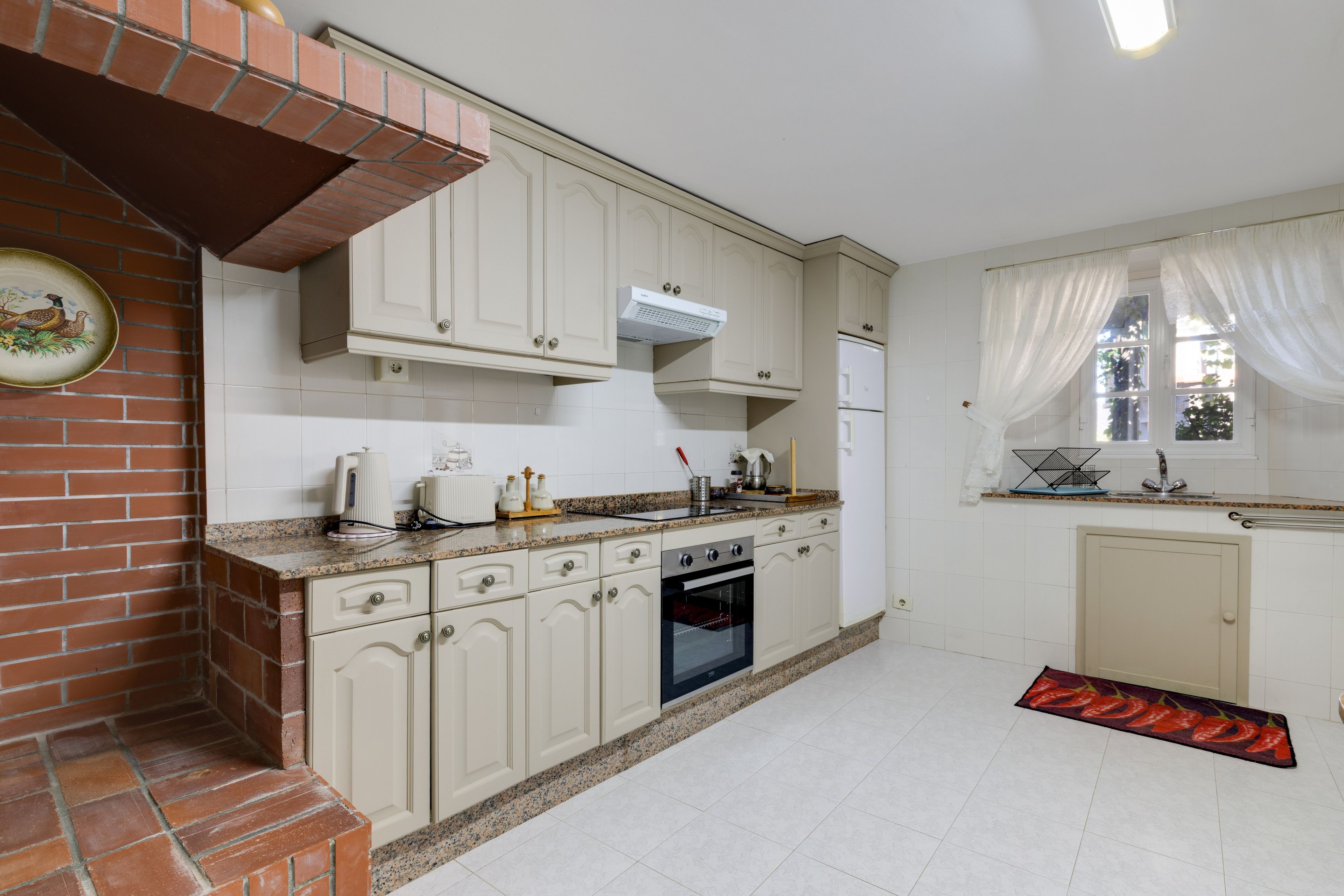 Una cocina con armarios blancos, una chimenea de ladrillo, una estufa y una ventana con cortinas blancas.
