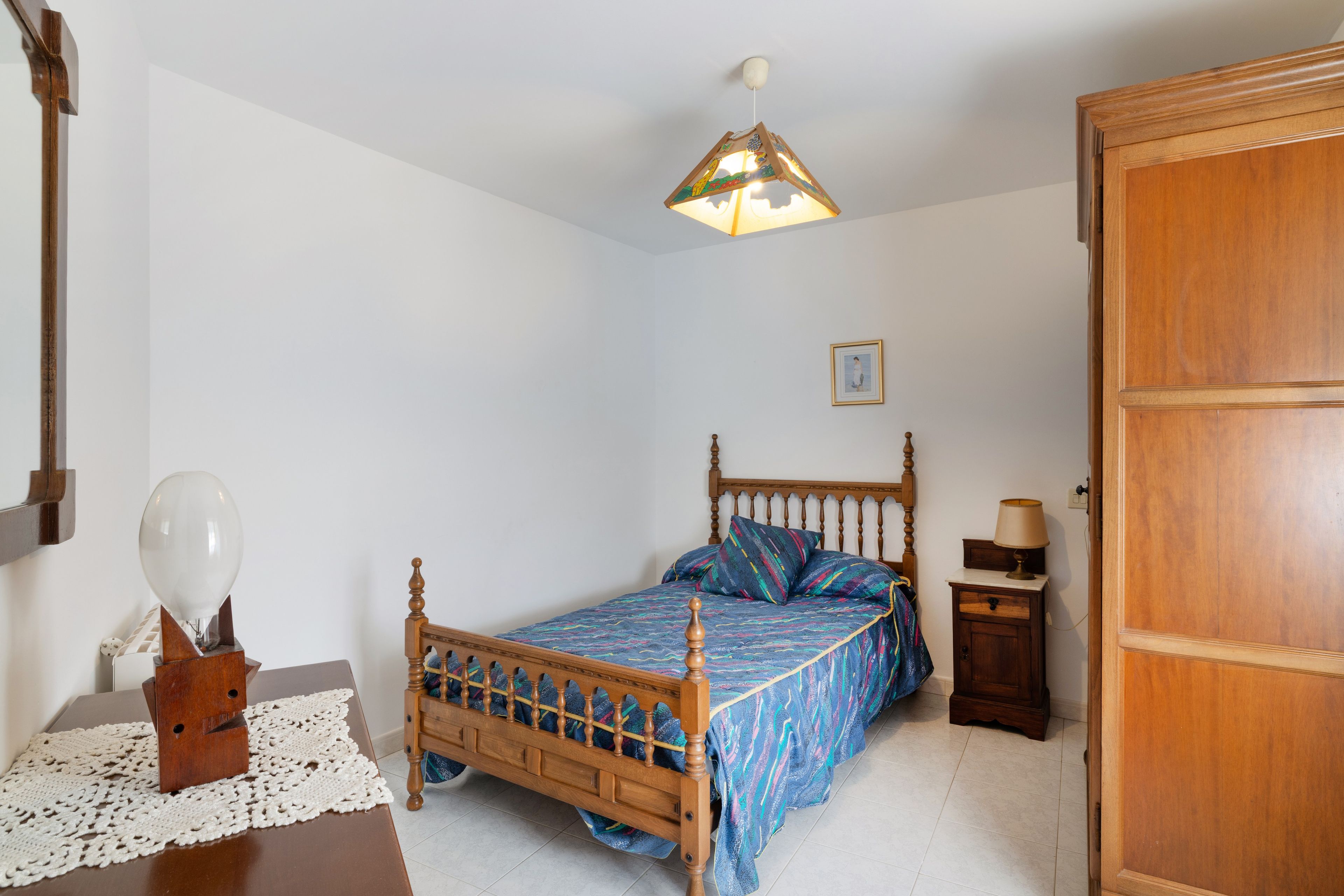 A bedroom with a wooden bed, a bedside table with a lamp, a wall-mounted light fixture, and a door.