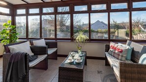 A sunroom with wicker furniture, a glass coffee table, and a view of a stone wall and buildings.