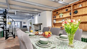 A dining area with a table set for four, surrounded by chairs, a bookshelf with decorative items, and a staircase in the background. - 3 Bed in St. Austell (oc-83541) (Gorran Haven)