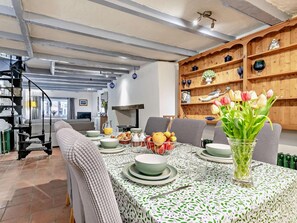 Un comedor con una mesa preparada para cuatro personas, rodeada de sillas, una estantería con objetos decorativos y una escalera al fondo.