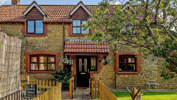 A stone building with a tiled roof, a wooden front porch, and a paved walkway leading to the entrance. - 3 Bed in Crewkerne (oc-83943) (Haselbury Plucknett)