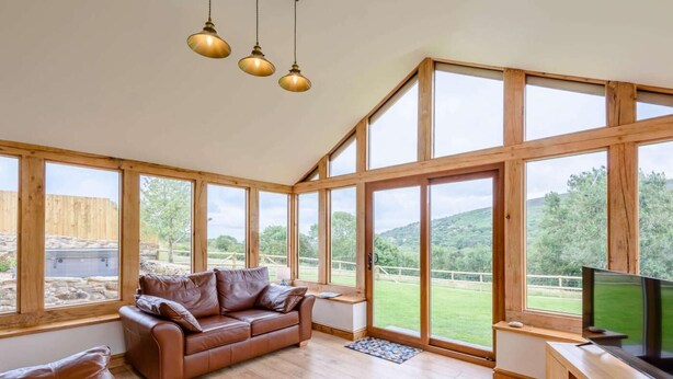 A spacious room with a large window, a leather sofa, a wooden TV stand, and a ceiling light fixture. - 3 Bed in Wrexham (oc-83938) (Coedpoeth)
