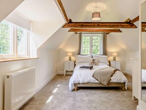 A bedroom with a sloped ceiling, a bed with white bedding, two bedside tables with lamps, a radiator, and a window with a view of greenery.
