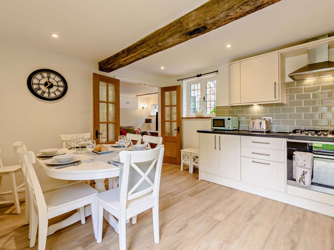Una cocina moderna con armarios blancos, una viga de techo de madera y una zona de comedor con sillas blancas y una mesa dispuesta para dos.