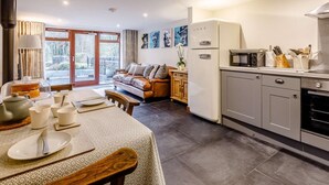 A modern kitchen with a refrigerator, microwave, and oven. There is a dining area with a table set for two, and a sofa in the background. - 1 Bed in Frome (oc-83834) (Holwell)