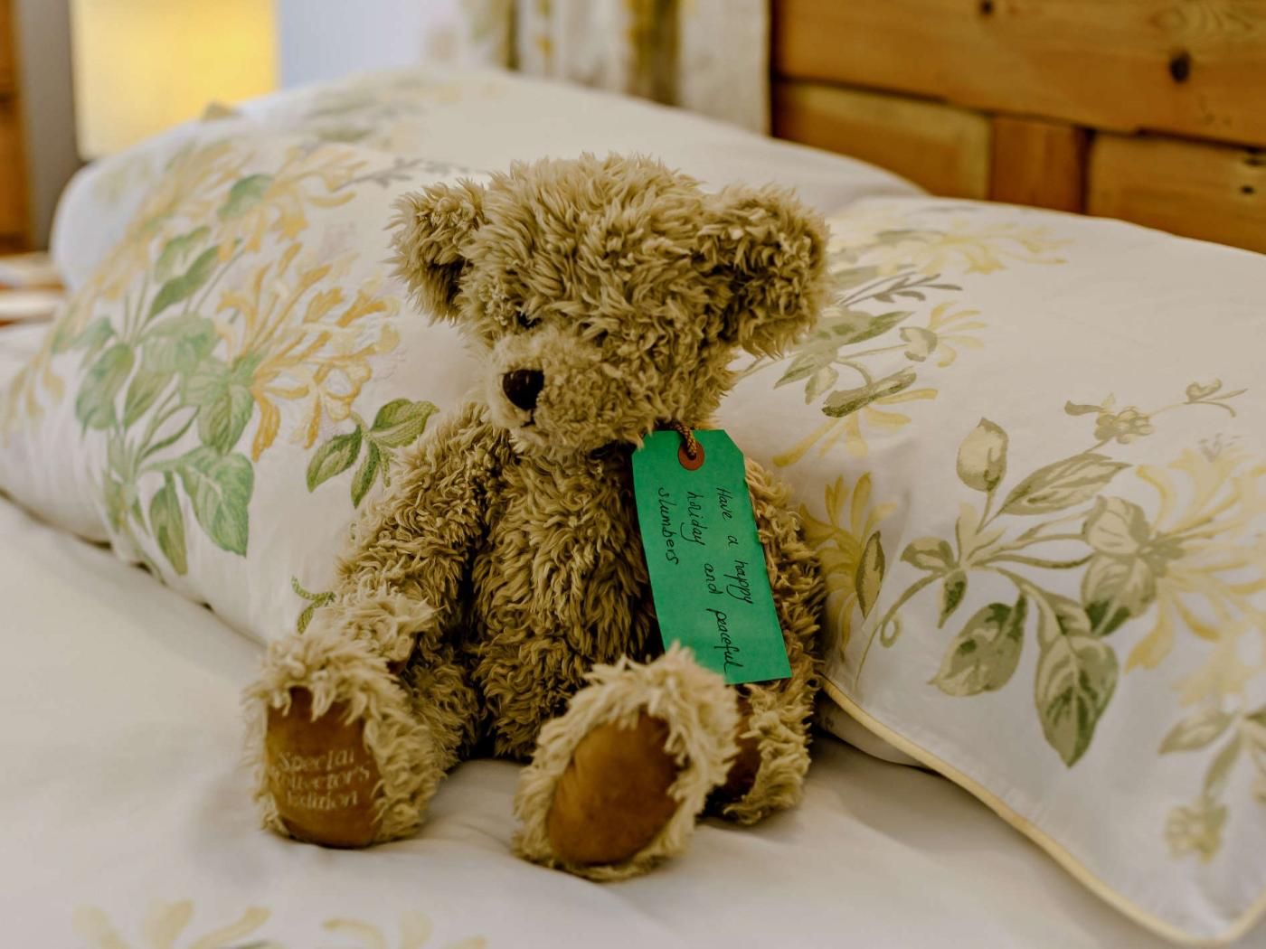 Un ours en peluche avec une étiquette, posé sur un lit recouvert d’une literie à motifs floraux.