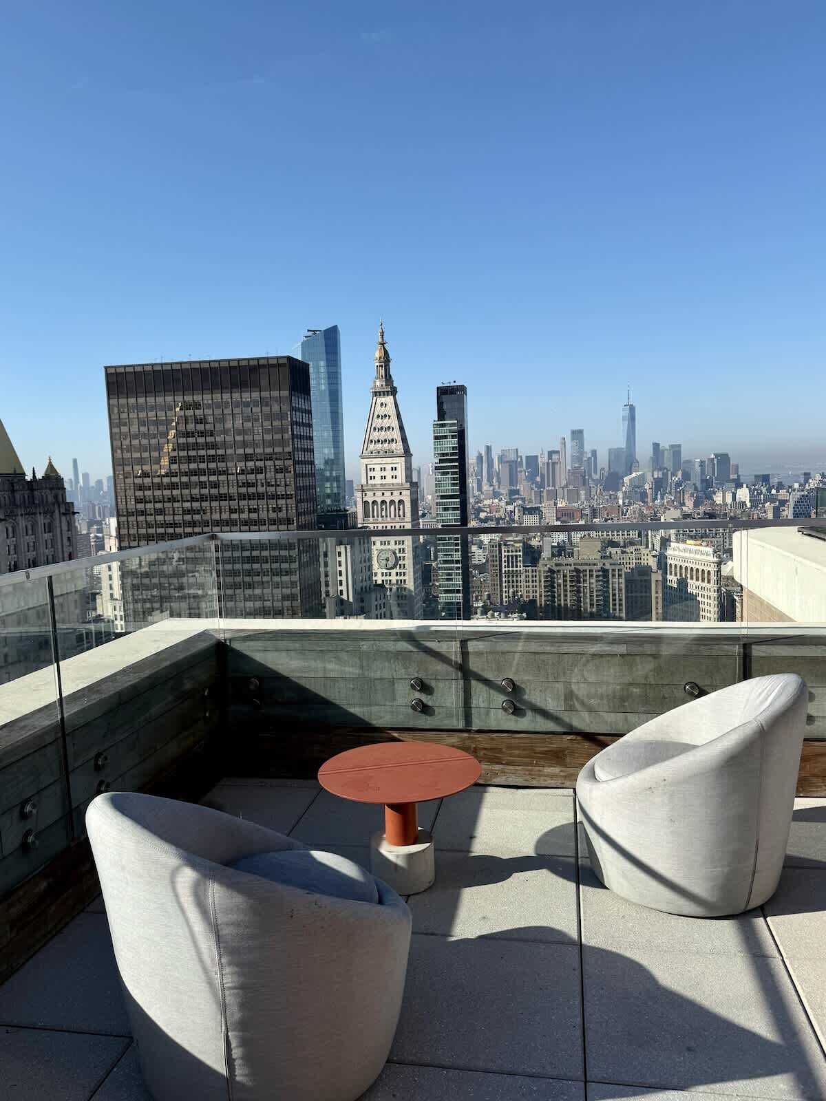 Eine Dachterrasse mit Blick auf die Stadtlandschaft, einer Glasumlaufung und zwei weißen Stühlen.