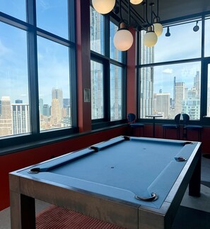 A pool table in a room with large windows, a city view, and hanging light fixtures. - Nomad Luxury Pied-a-Terre NYC (NY)